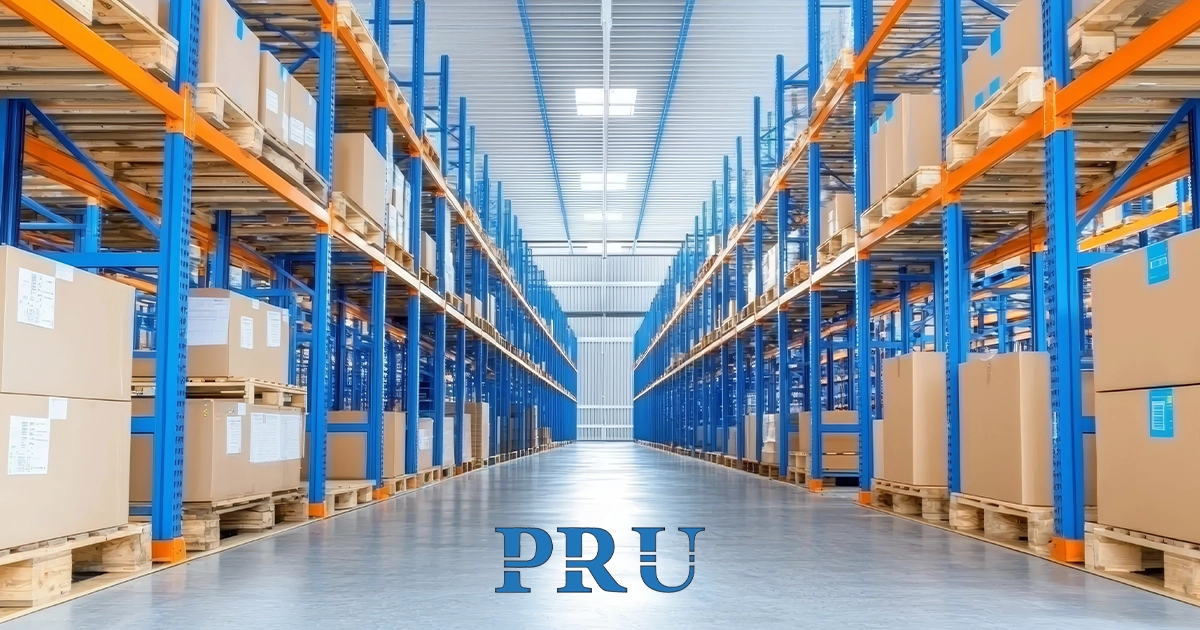 A large warehouse interior with long rows of blue and orange industrial storage racks filled with cardboard boxes on pallets.